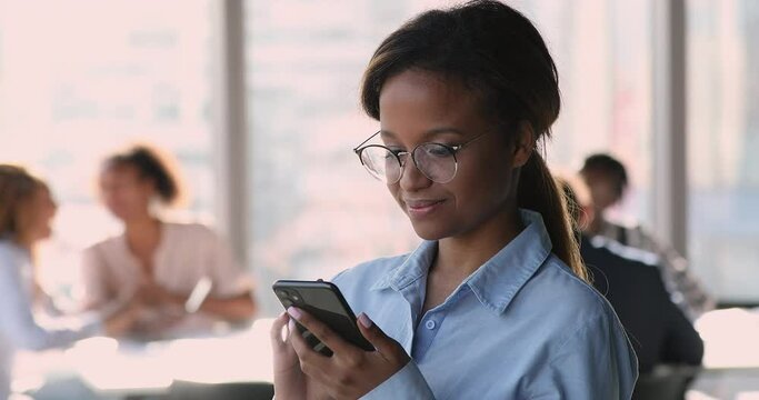 Attractive young African businesslady in glasses standing at workplace hold smartphone, using mobile version of web page with professional content, read news relevant to industry and interests concept