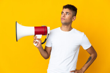 Young brazilian man isolated on yellow background holding a megaphone and thinking