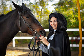 Fototapeta premium Happy girl in medieval clothes and a black cloak stands with a horse horse on the background of trees and a fence