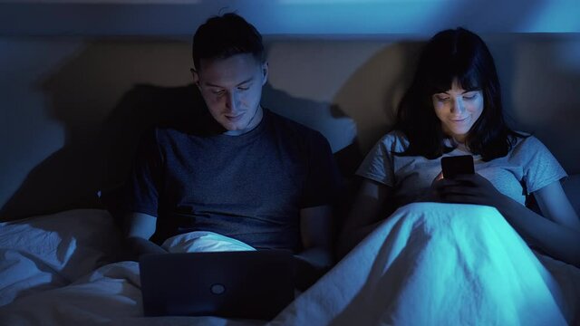 Gadget night. Digital couple. Modern technology. Amused woman enjoying using mobile phone man working online with laptop in bed in dark home bedroom with blue moonlight.