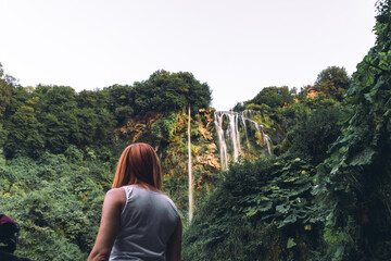 Naklejka premium Tourist girl pose, look at waterfall in Italy (Cascata delle Marmore) in Umbria. small, calm water pressure. Evening, sunset. Summer, bottom view