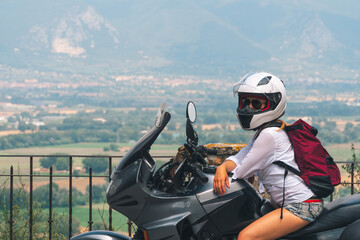 A lightly dressed girl is sitting on a motorcycle. No protection. Helmet. Sunny day in Narni, Italy. Green mountains in the background. Vacation and travel concept