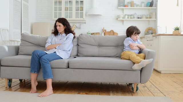 Annoyed mom and stubborn child sitting on couch at home, ignoring each other, posture of discontent. Sad mother disobedient kid son sitting apart on sofa with arms crossed, not talking after quarrel.