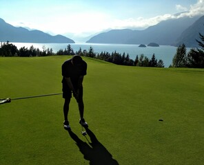 Man putting golf ball into hole with the ocean of Howe Sound British Columbia in the background.  Very scenic