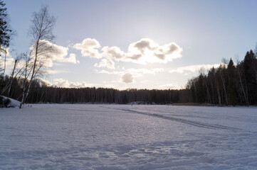 Beautiful winter (early spring)landscape with forest, frozen lake surface with pathway and the sun shining through the clouds.
