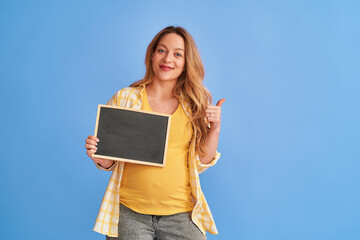 young, attractive, pregnant woman holds a board with inscriptions in her hands. Happy mom-to-be holds an empty ad banner, and points a thumbs up at the blue studio background, copy space
