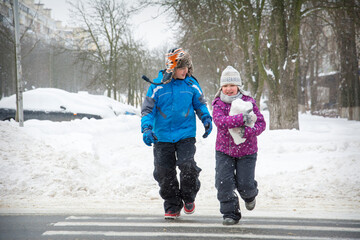 In winter, on the street in snowy weather, a boy and a girl cross the road along a pedestrian crossing.