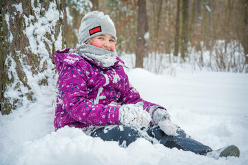 Fototapeta na wymiar In the winter afternoon in a snowy forest, a girl sits in the snow under a tree.
