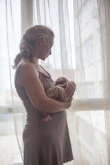 Young mother holding on hands  newborn two weeks baby at home in the room. Home portrait of  happy fаmily against the window