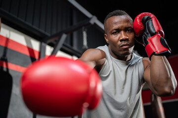African American fighter man wear boxing red gloves and punching forward with one fist to camera in the fitness sport gym. Make looking forward with serious face. Exercise for a healthy body