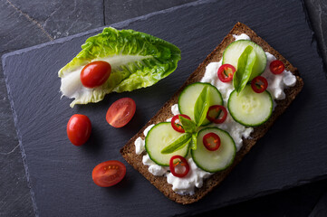 Fresh spring sandwich on a sliced gluten free sunflower bread. Red and green vegetables on black slate. Cucumber, chili and cottage cheese garnished with tomato and green leaves