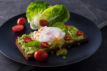 Fresh spring sandwich on a sliced gluten free sunflower bread. Red and green vegetables on black slate. Poached egg, avocado, chili and spring onion garnished with plum tomatoes