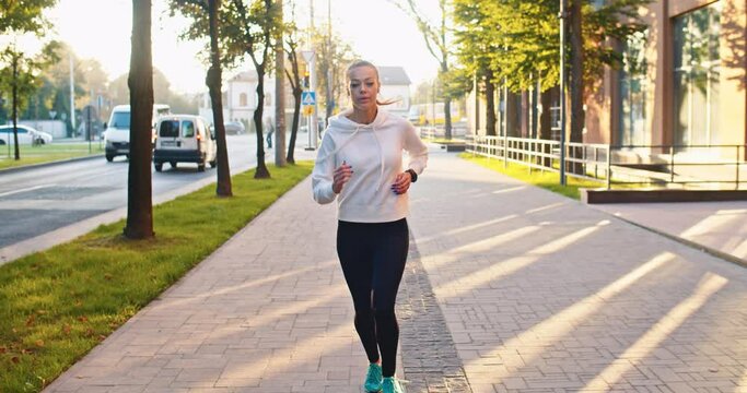 Front view of running Caucasian woman in sportswear doing cardio workout in street. Adult female athlete jogging, training before marathon competition in city. Health, success concept.