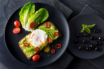 Fresh spring sandwich on a sliced gluten free sunflower bread. Red and green vegetables on black slate. Poached egg, avocado, chili and spring onion garnished with plum tomatoes  and black olives