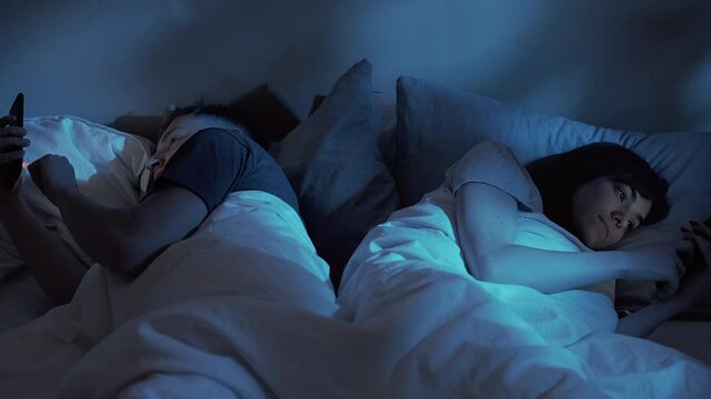 Gadget night. Digital couple. Family communication problem. Social media addiction. Bored husband wife using mobile phone phubbing ignoring each other in bed late in dark home bedroom.