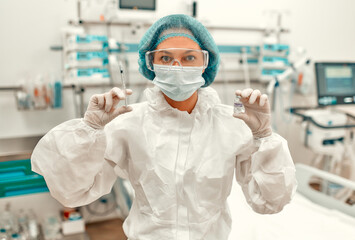 A doctor in a protective suit holds a syringe and an ampoule with the covid-19 coronavirus vaccine while standing in an intensive care unit in a modern hospital.