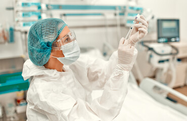 Fototapeta premium A doctor in a protective suit holds a syringe and an ampoule with the covid-19 coronavirus vaccine while standing in an intensive care unit in a modern hospital.