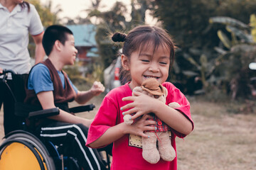 Cute Asian girl holds a teddy bear doll playing and smiling face with  blurry background of Asian special child on the outdoor park, Disable people family concept.