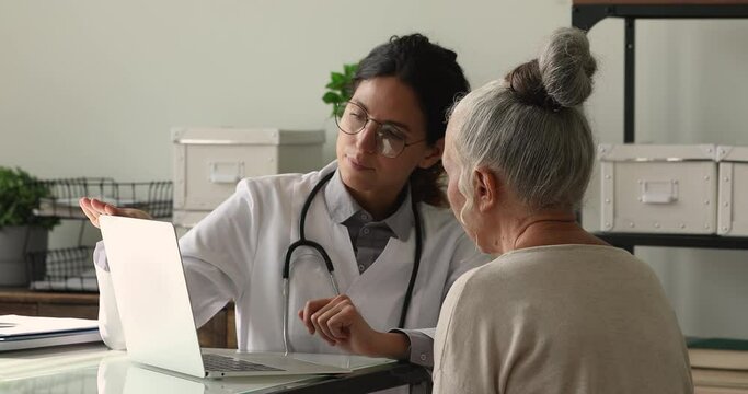 Professional young 35s female physician showing medical insurance presentation or health test results on laptop to interested old mature retired patient at clinic meeting, discussing illness treatment