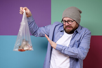 Man holds a smelly, spoiled fish in a transparent bag, closes eyes because of the unpleasant smell and makes a stop gesture.  - obrazy, fototapety, plakaty