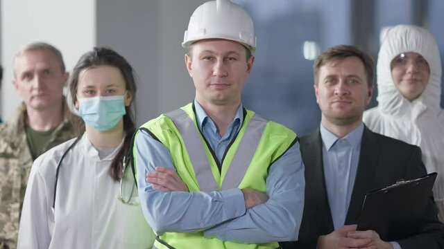 Confident smiling male builder looking at camera crossing hands standing indoors with group of people of different professions. Portrait of Caucasian man in hard hat posing. Construction industry