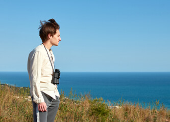 A side view of an attractive young man with binoculars hanging around his neck in front of lovely sea scenery on a windy day