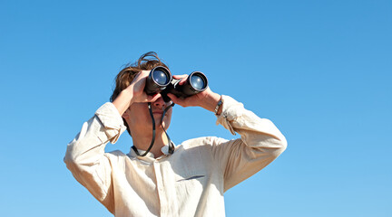 Obraz premium A young caucasian male looking at a view with binoculars under the bright blue sky