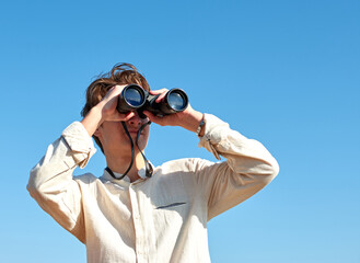 Obraz premium A young caucasian male looking at a view with binoculars under the bright blue sky