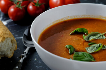 Close-up of a bowl with tomato soup and basil, next to it a baguette.