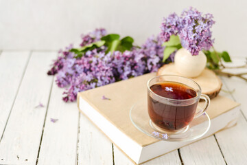 Romantic background with a cup of tea, lilac flowers and a book over a white wooden table. Leisure concept, spring breakfast.