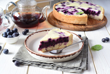 Delicious blueberry cake and a slice of blueberry cake on a white plate. A piece of berry pie on a nice plate and a cup of hot tea on a white table.
