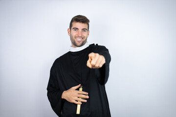 Young hispanic man wearing priest uniform standing over white background laughing at you, pointing finger to the camera with hand over body, shame expression