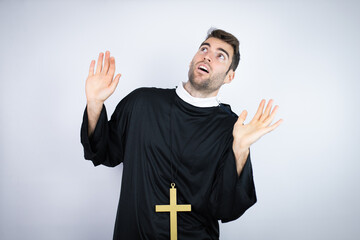 Young hispanic man wearing priest uniform standing over white background scared with her arms up...