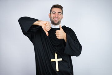Young hispanic man wearing priest uniform standing over white background Doing thumbs up and down,...