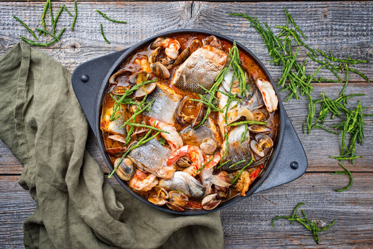 Modern style traditional Spanish seafood zarzuela de pescado with fish, king prawns and venus clams served with sea asparagus in red sauce as top view in design pot