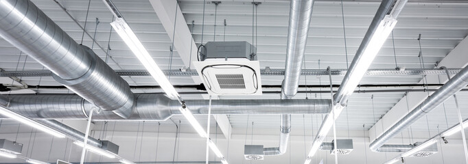 Ceiling mounted cassette type air condition units with other parts of ventilation system (tubes, cables and vents) located inside commercial hall with hanging lights and other construction parts. - obrazy, fototapety, plakaty