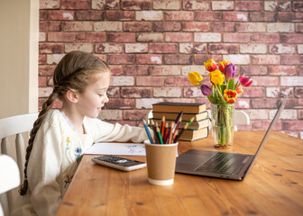 School closed cute pretty young girl doing work writing calculations at home school with computer laptop calculator pot of pencils on colourful desk with flowers happy and smiling whilst working hard