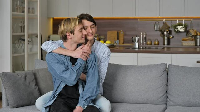 Homosexual guys. Loving gay couple, sitting at home, tenderly hugging and caressing each other, relaxing together on the sofa in the living room, spend time together, same-sex couple