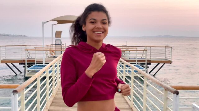 Keep going. Close up of smiling young woman jogging along the beach on the wooden pier at dawn. Slow motion. Healthy lifestyle, nature concept