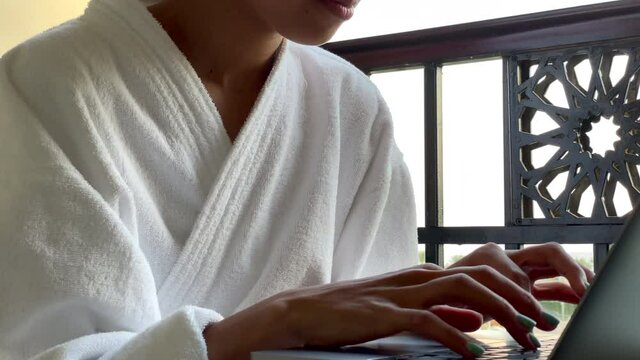 Remote work. Young woman in bathrobe working on the laptop while sitting on the hotel balcony during vacation. Freelance, relax, summertime concept