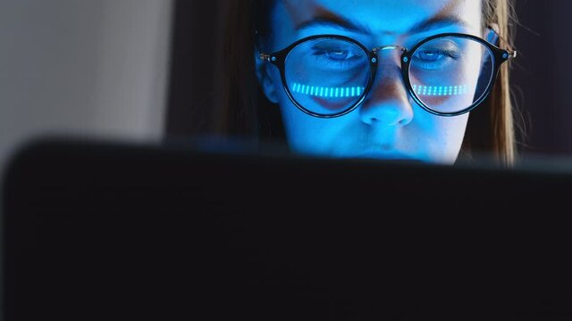 Focused young millennial woman in glasses look at laptop screen pondering considering making notes, typing studying e learning on pc technology sit on bed at home. Distance online education concept