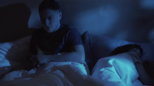Cheating husband. Night infidelity. Marital disloyalty. Sneaky man hiding phone from sleeping wife enjoying chatting with secret lover in bed late in dark home bedroom.
