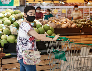 Asian 40s woman wearing face mask  push shopping cart in suppermarket departmentstore. Girl choosing, looking grocery things to buy at shelf during coronavirus crisis or covid19 outbreak.
