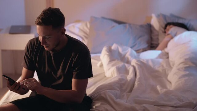 Night cheating. Secret affair. Husband betrayal. Sneaky man hiding phone from sleeping wife enjoying using dating app sitting on bed late in home bedroom.