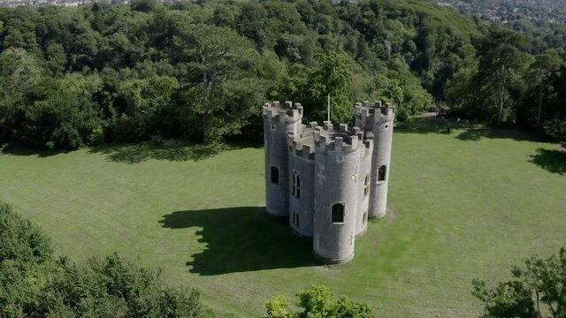Aerial move around 18th century gothic British folly castle with three turrets on a sunny day with green woodland in the background