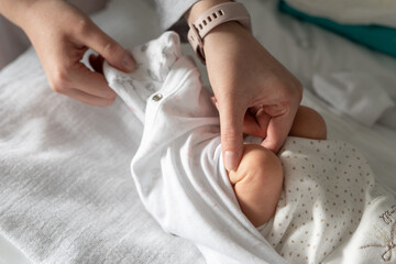 Close up on hand of unknown woman holding leg of her newborn baby - caucasian mother dressing or undressing her little baby on the bed at home in day - motherhood and growing up concept