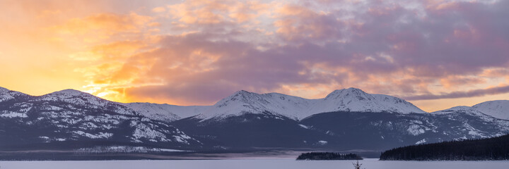 Fototapeta na wymiar Panorama of stunning sunset pink, orange, yellow view with snow capped hige mountains, frozen lake, wilderness, forest and woods in northern Canada. 