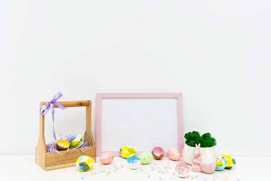 Front view of Easter holiday composition. Hand painted colorful eggs in trendy colors, basket, geometric vase with moss, confetti in eggshell, pink Easter bunny. Photo frame mockup for Easter design