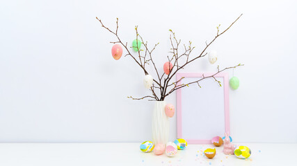 Trendy colorful Easter concept with hand painted eggs in yellow, grey colors, geometric vase, confetti, twigs, pink Easter bunny in light interior. Blank photo frame, mockup with copy space for text.