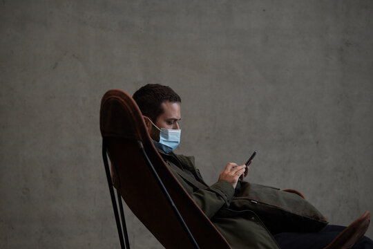 Young man with face mask sitting on chair with mobile phone
